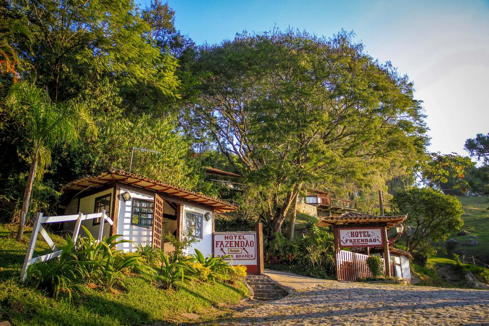 Entrada do Hotel Fazendão ao entardecer, com casinha de recepção, placas e jardim