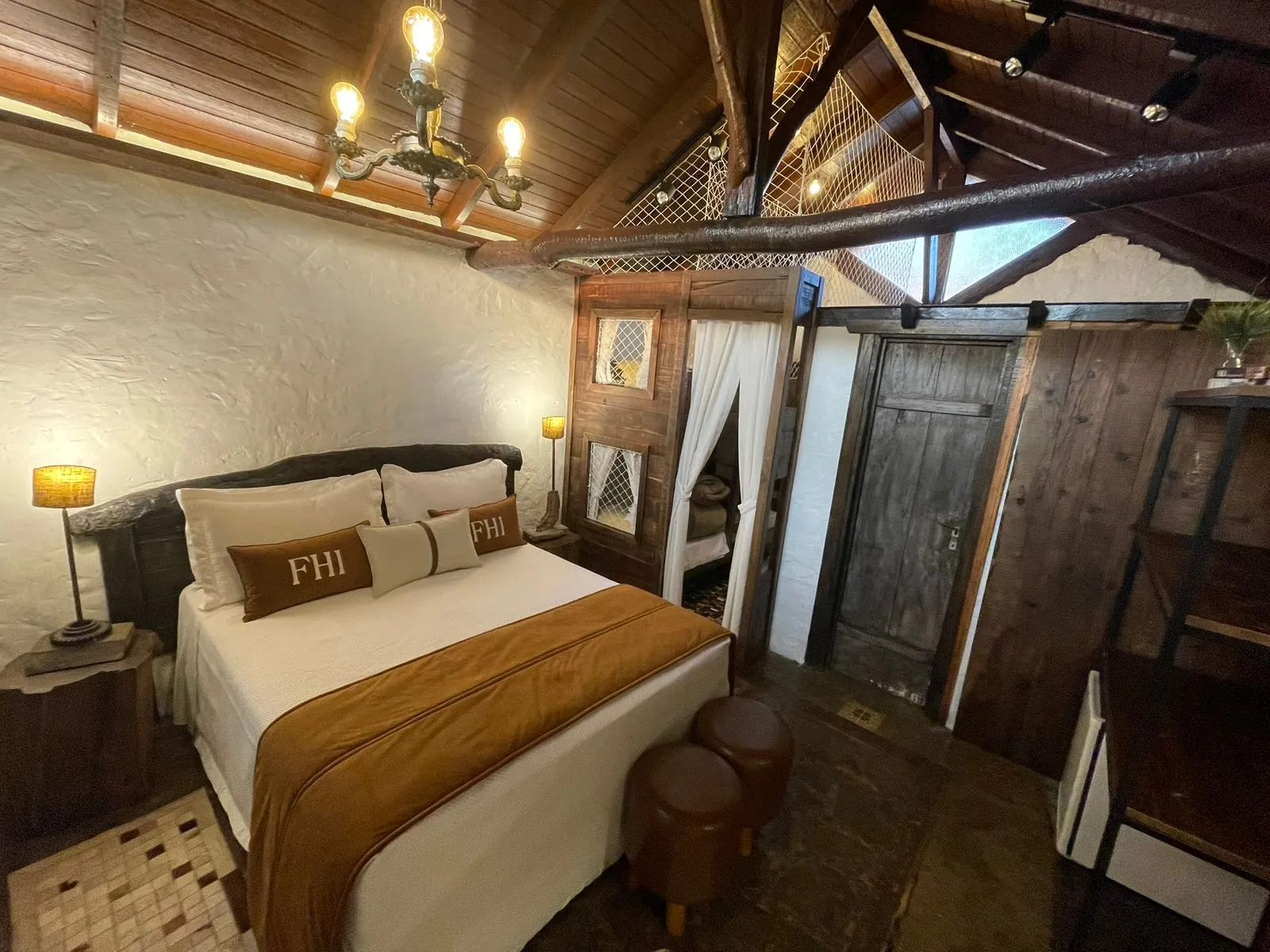 Quarto rústico com cama de casal, lustre e travesseiros com o monograma FHI no Fazenda Hotel Invernadinha