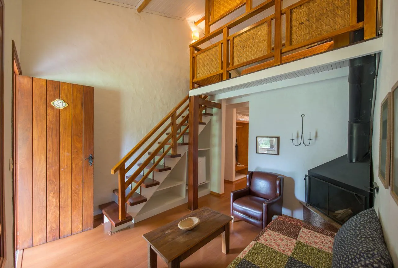 Sala de estar do chalé Colônia com mezanino, lareira e escada de madeira