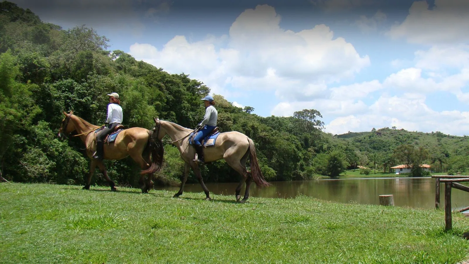 Cavalgada de hóspedes às margens do lago, com a mata e chalés ao fundo